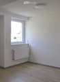 Empty room with wooden flooring, white walls, a window, a radiator, and a door. No furniture or equipment is present.