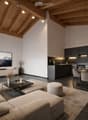 Open-concept living and dining area with a modern aesthetic. Features a large sectional sofa, coffee table, ottoman, dining table with chairs, and a sleek kitchen with dark cabinets and a central island. A large television is mounted on the wall, and the