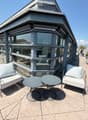 Modern rooftop terrace with a sofa, two round tables, and several chairs, surrounded by glass walls and a cityscape view.