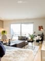 Modern open-plan living and dining area with a grey sofa, coffee table, armchair, ottoman, floor lamp, dining table with chairs, kitchen cabinet, faucet, plants, picture frame, bottle, and wine glass.