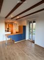 Bright, empty room with wooden flooring, white walls, and exposed wooden beams on the ceiling. Features a sliding glass door leading to an outdoor area and a small kitchen area visible in the background.