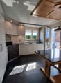 Modern kitchen with light-colored cabinets, a central island, and a dining area with a black countertop table and wooden chairs. The space features large windows and a pendant light hanging from the ceiling.
