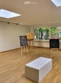 An art studio with wooden floors, large windows, and several easels displaying paintings. A white stool is placed in the center of the room.