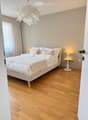 Modern bedroom with a large bed, white bedding, wooden floor, nightstand with lamp, and sheer curtains.