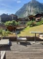 Wooden deck with outdoor furniture including a table, chairs, and a bench, overlooking a scenic mountain view with surrounding houses and greenery.