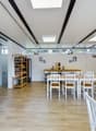 Bright, modern room with wooden flooring, white walls, and slanted ceiling. Features include a long wooden counter, several tables and chairs, a shelf with bottles, and a refrigerator.