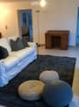 Modern living room with a white sofa, decorative pillows, a dark gray rug, three round woven stools, a small black side table, a white TV stand, and a dining area with chairs and a table visible in the background.