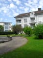 Well-maintained garden area with a circular stone fountain, neatly trimmed hedges, and a paved walkway leading to a multi-story residential building with balconies.