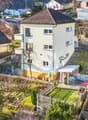 Aerial view of a two-story house with a sloped roof, surrounded by a garden and outdoor seating area. The house has multiple windows and a small staircase leading to the entrance. The garden includes a patio with a table and chairs, and there are addition