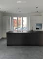 Modern kitchen with grey cabinets, a central island, and large windows overlooking a street with parked cars.