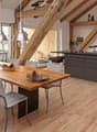 Open-plan kitchen and dining area with light wooden flooring, exposed wooden beams, modern kitchen island with cabinets, dining table with chairs, pendant lights, and decorative bowl on the table.