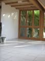 Bright, empty room with white tiled flooring, large wooden-framed windows, and a modern black wood-burning stove.