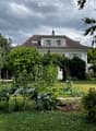 A well-maintained garden with a variety of plants and vegetables, featuring a white house with a red-tiled roof in the background.