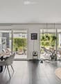 Modern dining area with a wooden table, grey chairs, pendant lights, shelves with bottles, a vase with flowers, and a cat tree. Large glass doors open to a patio with outdoor furniture and plants.