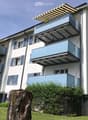 Modern multi-story residential building with white facade, blue shutters, and multiple balconies with glass railings and retractable awnings.