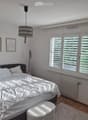 Bright bedroom with a large bed, white bedding, a small stool, radiator, and three picture frames on the wall. Large windows with blinds let in natural light.