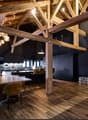Open-concept dining and kitchen area with exposed wooden beams, wooden flooring, and modern lighting. Features a large wooden dining table with chairs, a kitchen island with stools, pendant lights, and a large picture frame on the wall.