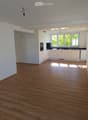 Empty room with wooden flooring and white walls, leading to a small kitchen area with white cabinets and black countertops.