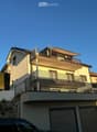 Modern two-story house with balconies, light yellow facade, and multiple windows. Features include a fenced yard, garage, and a raised ground floor.