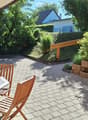 A sunny patio with a wooden table and four chairs, surrounded by greenery and a brick wall.
