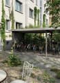 Garden area with stone-paved ground, metal chairs, a round table, bicycles under a covered area, and a building with white walls and large windows.