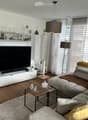 Modern living room with a grey sectional sofa, wooden coffee table, TV stand, cat tree, and decorative shelves. The room features large windows with sheer curtains and a pendant light hanging from the ceiling.