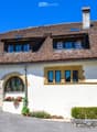 A two-story house with a tiled roof, white exterior walls, and multiple windows. The house features an arched entrance with potted plants and flowers, and is surrounded by well-maintained greenery and shrubs.