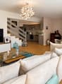Modern living room with a white sofa, glass coffee table, and a TV stand. The room features a staircase leading to an open-plan kitchen area with modern appliances.