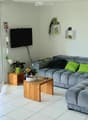 Bright living room with large sliding glass doors opening to a garden, grey sectional sofa with green cushions, wooden coffee table, cat tree, cat house, small table with flower pot, and a television mounted on the wall.
