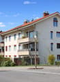 A multi-story residential building with a red-tiled roof, white walls, multiple windows, and balconies. The building is surrounded by greenery and a street with traffic markings.