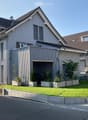 Modern two-story house with grey siding, multiple windows, and a garage entrance. The front yard features a well-maintained lawn, potted plants, and a small garden area.