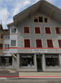 3 story building, gray exterior, red shutters, storefronts on the ground floor, sign 'wenger wohnen', sign 'Polsterarbeiten', sign 'Vorhänge Teppiche'