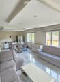Bright living room with a grey sectional sofa, coffee table, dining area with chairs and table, kitchen area with refrigerator and cabinets, large windows with blinds, and a TV on the wall.