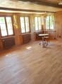 Empty room with wooden flooring, wooden walls, large windows with yellow curtains, radiators, and a small round table with a chair near the windows.