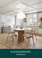 Bright, modern living and dining area with wooden flooring, large windows, a round dining table with four chairs, a sofa, a pendant light, a fireplace with stacked logs, and a potted plant.