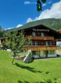 A two-story chalet with a balcony, surrounded by a well-maintained garden and a mountain backdrop.