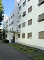 Modern multi-story building with white facade, multiple windows, and a small garden area with flowers and a tree.