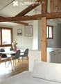 Bright, open-plan living and dining area with exposed wooden beams, large windows, a sofa, dining table with chairs, and a small kitchen area with cabinets and a refrigerator.