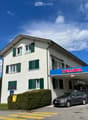 Two-story white building with green shutters, Tamoil gas station attached, several parked cars, and a clear blue sky.