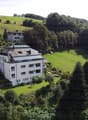Aerial view of a modern white building with a flat roof and large windows, surrounded by lush green hills and trees. The building has a Swiss flag displayed in front.