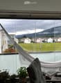 Balcony with large windows offering a scenic view of a green field, mountains, and buildings. Features a hanging chair, a small table, potted plants, and a hammock.