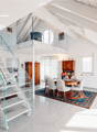 Modern, open-concept living and dining area with high ceilings, exposed wooden beams, and a glass staircase leading to an upper level. The dining area features a round wooden table with six upholstered chairs, a colorful area rug, and a vase with flowers