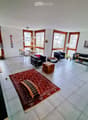 Bright living room with large windows, tiled floor, red rug, sofa, coffee table, cat tree, dining table, and chairs.