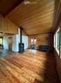 Spacious living room with wooden floors, exposed wooden ceiling beams, and a central fireplace. Features a dark green sofa and matching armchair, with large windows and a door leading outside.
