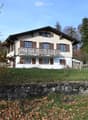Two-story house with a balcony, wooden shutters, and a stone retaining wall in front.