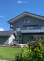 Two-story modern house with grey exterior, white trim, multiple windows, and a balcony on the upper level. The house features a well-maintained lawn and surrounding greenery.