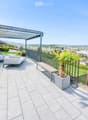 Modern rooftop terrace with a panoramic city view, featuring a sofa, a bench, and several potted plants, including palm trees and flowering shrubs.