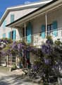 Building. Two-story house with grey stone facade, teal shutters, and a covered balcony adorned with blooming wisteria. The entrance features a bicycle and potted plants.