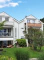 A two-story white house with balconies, surrounded by a well-maintained garden with outdoor furniture and greenery.