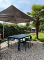 Garden with a table, chairs, a hammock, and a large umbrella, surrounded by greenery and a wooden stove.